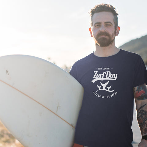 Junger Mann mit Surfbrett unter dem Arm trägt ein T-Shirt von Zurfday aus der SOS-Kollektion