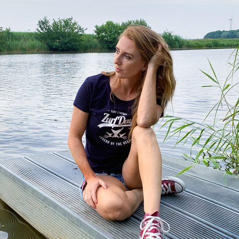 Eine junge Frau sitzt auf einem Steg an einem See und trägt ein Zurfday T-Shirt