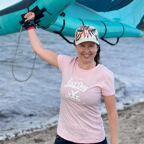 Eine junge Frau hält einen Kite Schirm, sie lächelt in dei Kamera und tragt ein rosa farbenes Zurfday T-Shirt