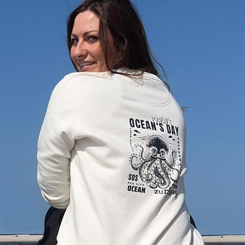 Eine junge Frau sitzt am Strand und schaut über die Schulter, sie trägt das Sweatshirt zum Ocean's Day