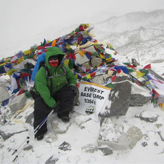 Plus size woman wearing a green jacket and black plants sitting on a pile of rocks and Buddhist prayer flags next to a sign that reads "Everest Base Camp 5364 m".