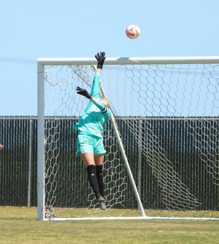 Teenage soccer player and goalkeeper in a blue jersey dives mid air to push the ball over the crossbar in a soccer game.