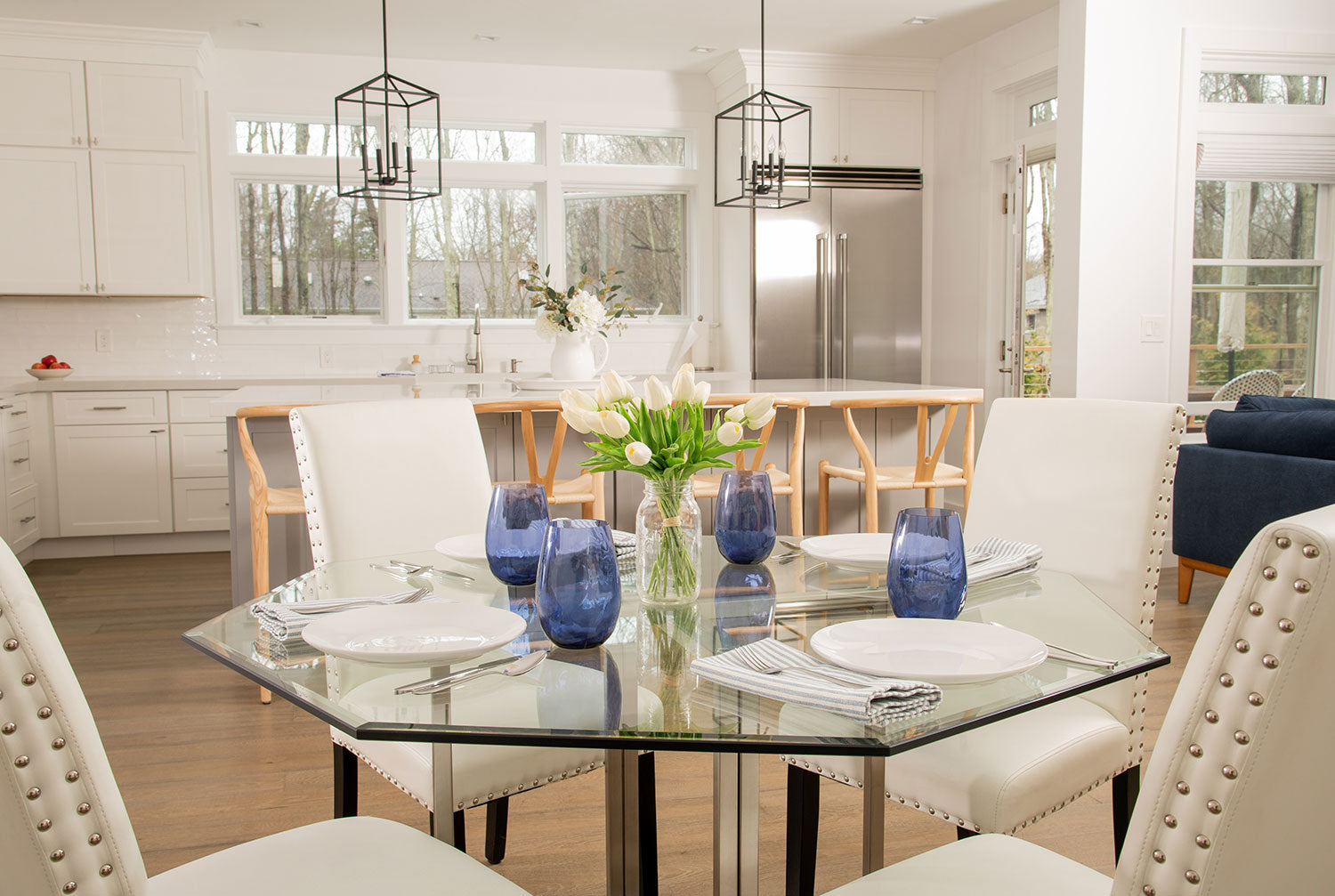 Octagon shaped clear glass table with white chairs and blue cylindrical cups on top next to kitchen island