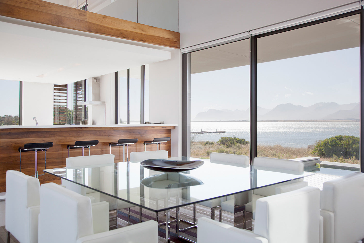 Square Glass Table with eight white chairs in the foreground and glass panes with a body of water in the background