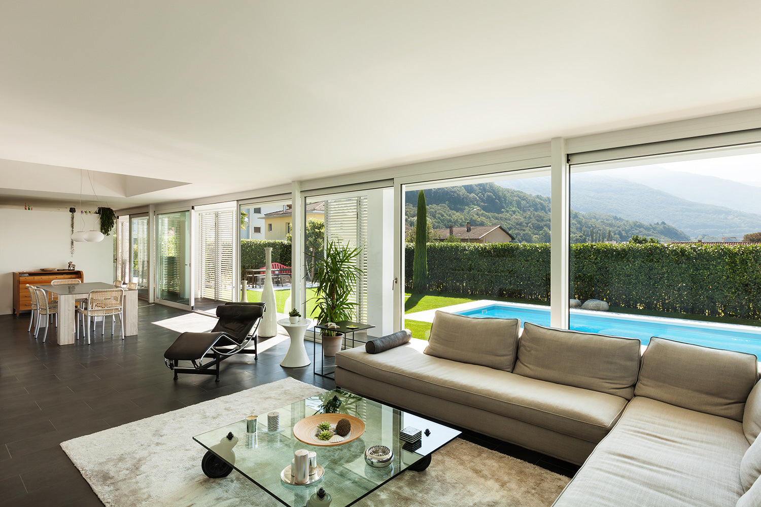 Square clear glass table next to beige sectional with glass panes behind it showing a rectangular pool outside