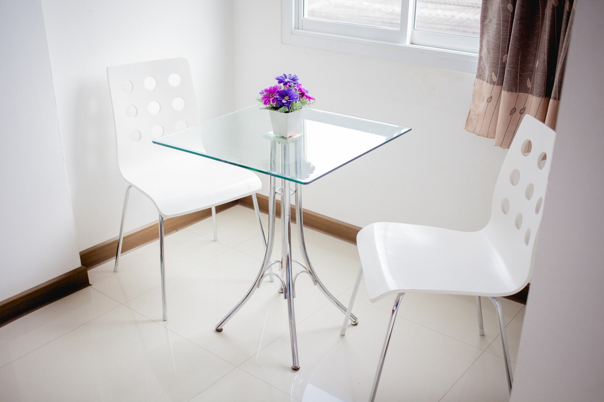 Square clear glass table top on steel base with two white chairs in corner of room