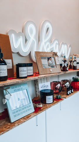 An urban ivy aromas logo with neon sign in the background of a shop display with things like a picture frame, candles, wax melts, hand soap and autumn leaves to decorate the shelves