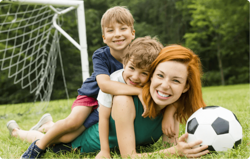 Two smiling kids are piled on top of their mother’s back while on the playground. The mother is holding a soccer ball.