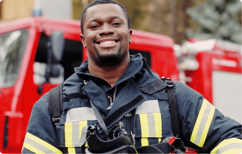 A fireman wearing a fireman suit is holding his hard hat while smiling.