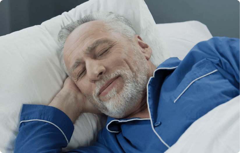A senior man wearing blue sleepwear is sleeping with a hint of a tiny smile on his lips.