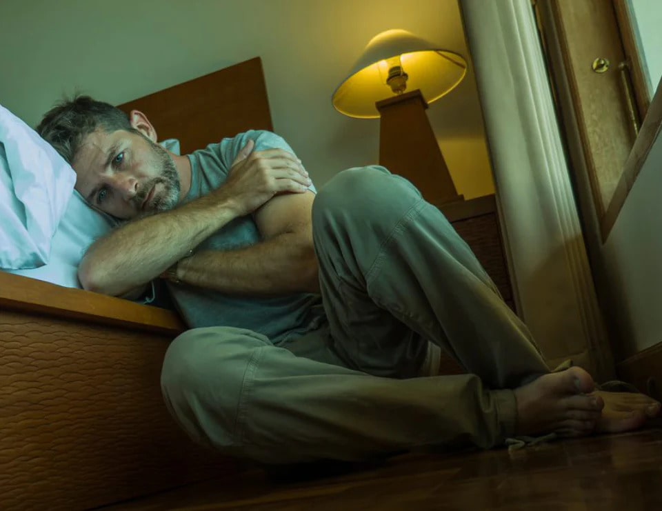 A man is sitting on the floor, his head leaning towards his bed with his arms crossed hugging himself.