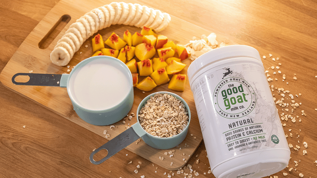 Sliced banana, 1 cup of goat milk, 1/2 cup oats, chopped nectarine and a container of Good Goat Milk powder on a wooden cutting board. 