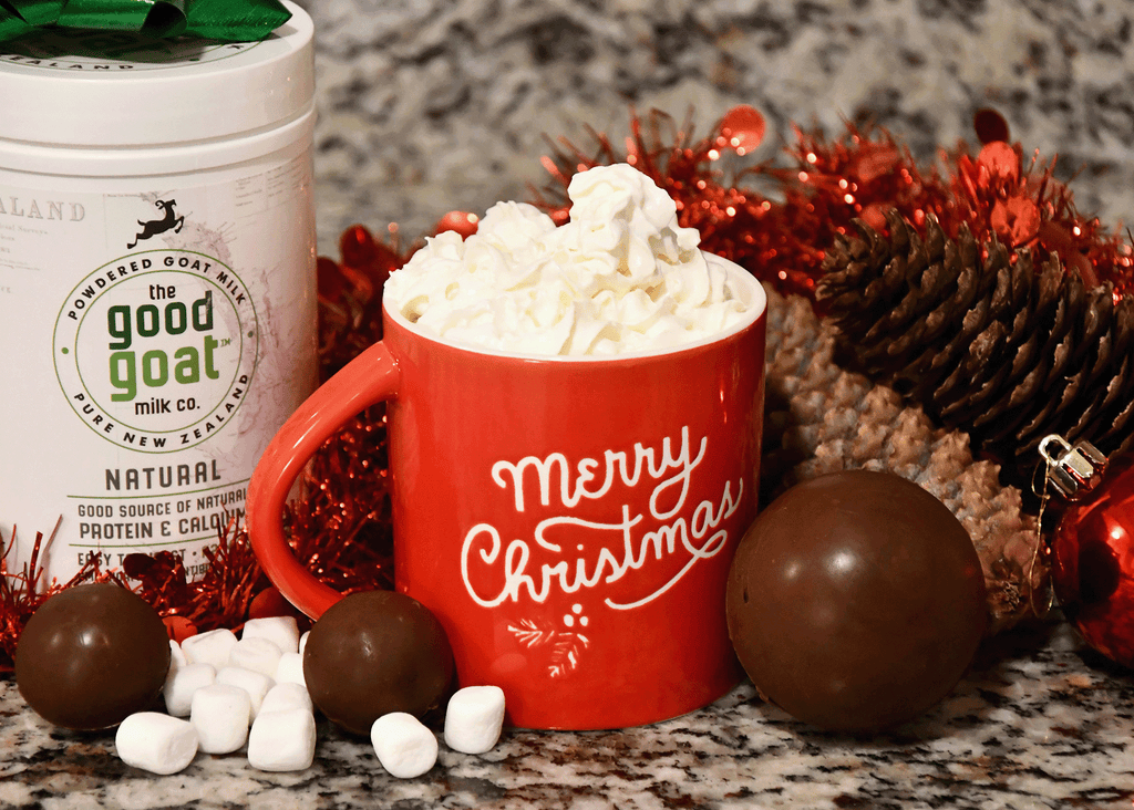 A red Merry Christmas mug with hot chocolate inside a lot of whip cream on top. Marshmallows, more chocolate bombs, pine cones, a red Christmas bobble and a container of Good Goat Milk Powder surrounding the mug.