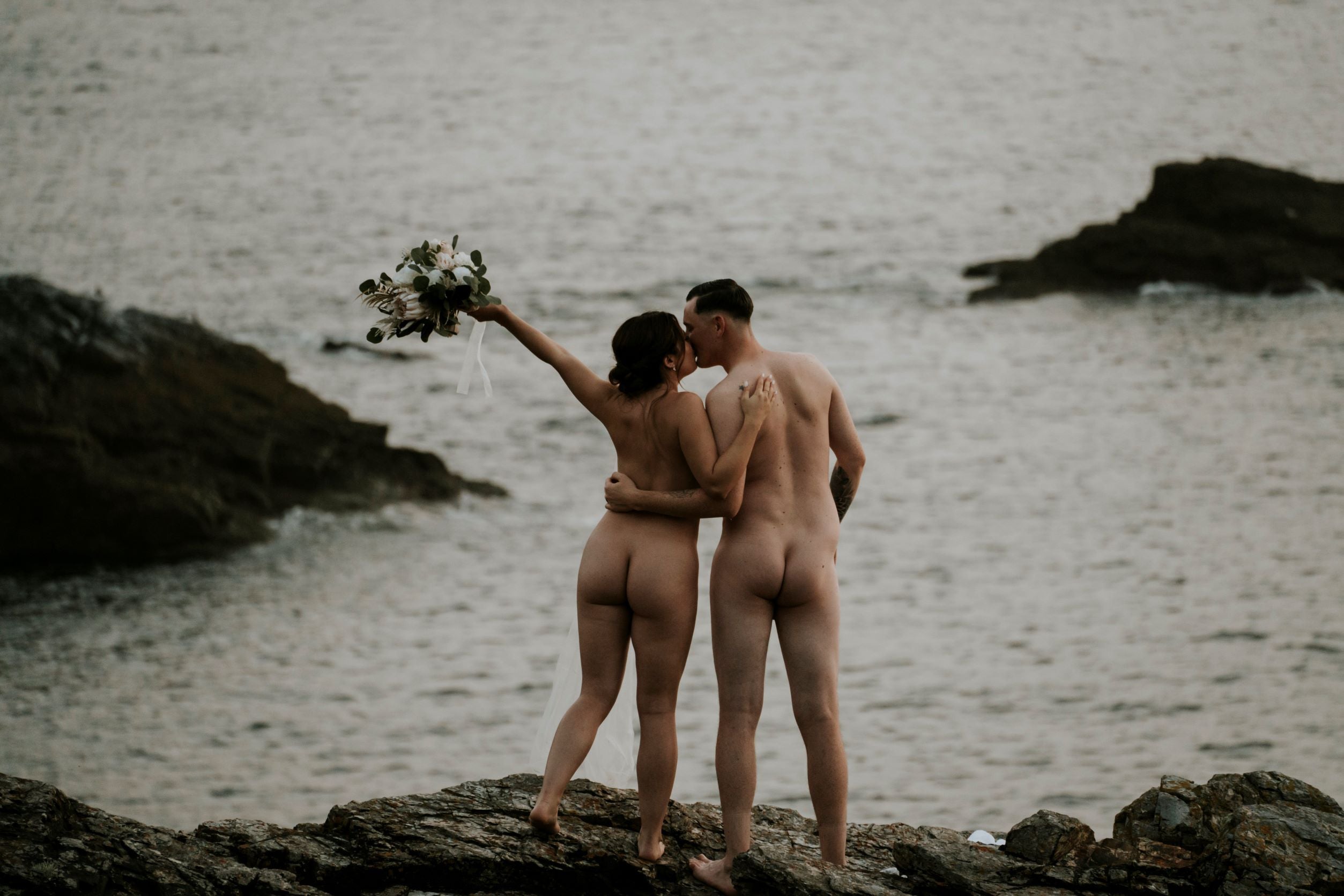 Nude couple with bouquet kiss within a seascape  