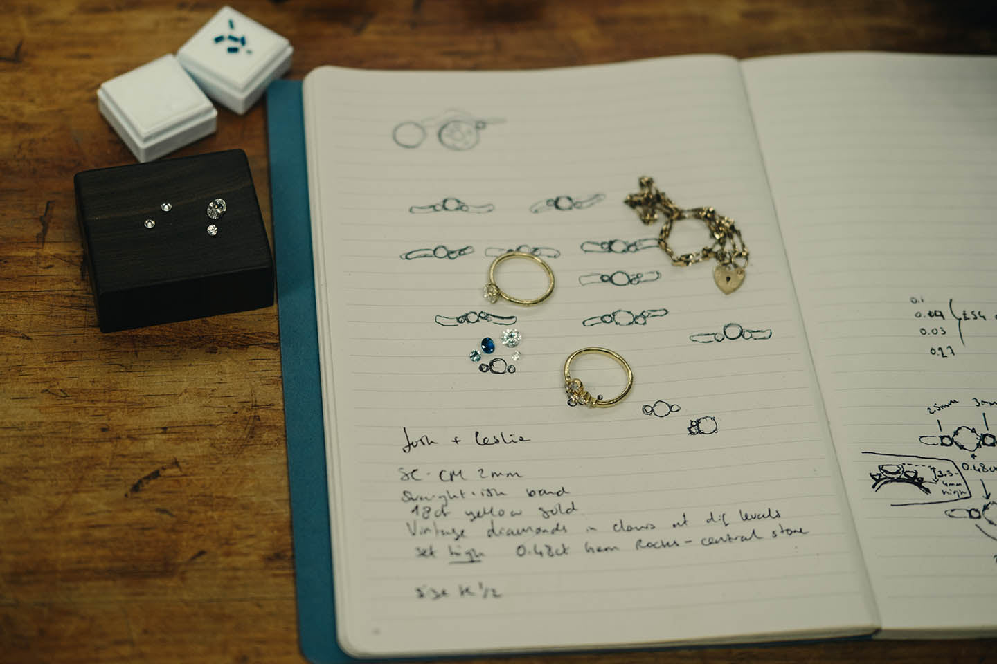 The process of commissioning a piece of jewellery - a notebook lays open with drawings and small annotations alonside examples of rings and loose gemstones and diamonds