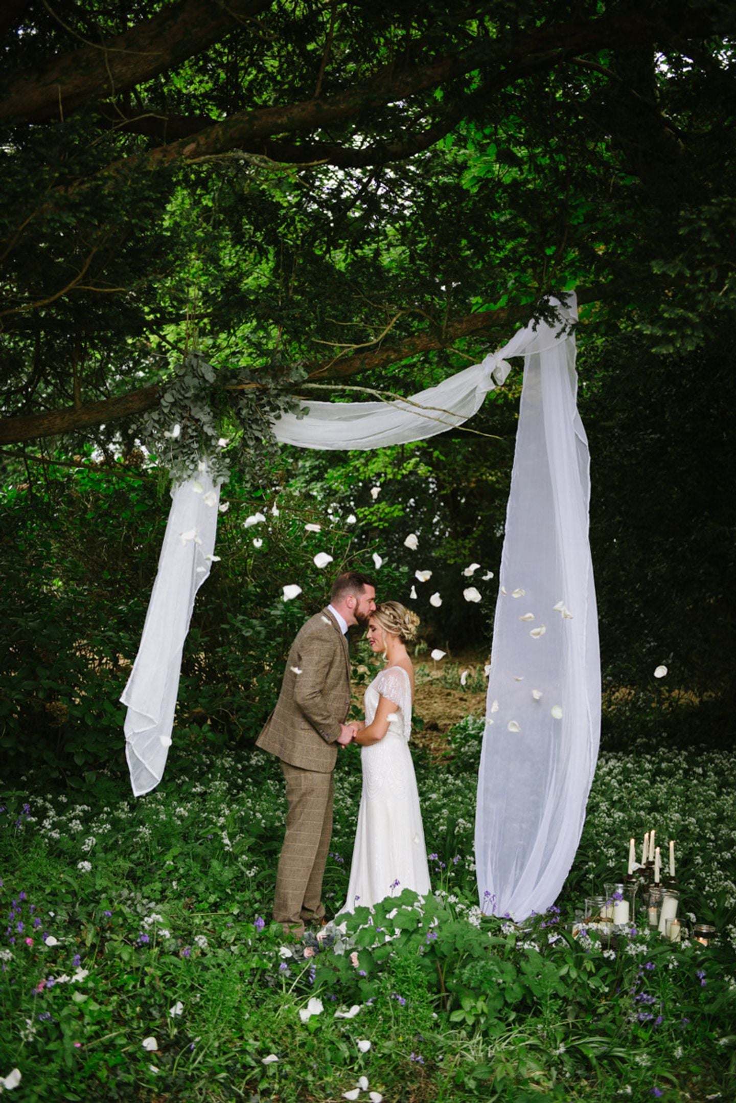 Simple woodland wedding ceremony styling, bride and groom stand below minimal white drapes under trees, he kisses her head, candles are on the floor and rose petals float by