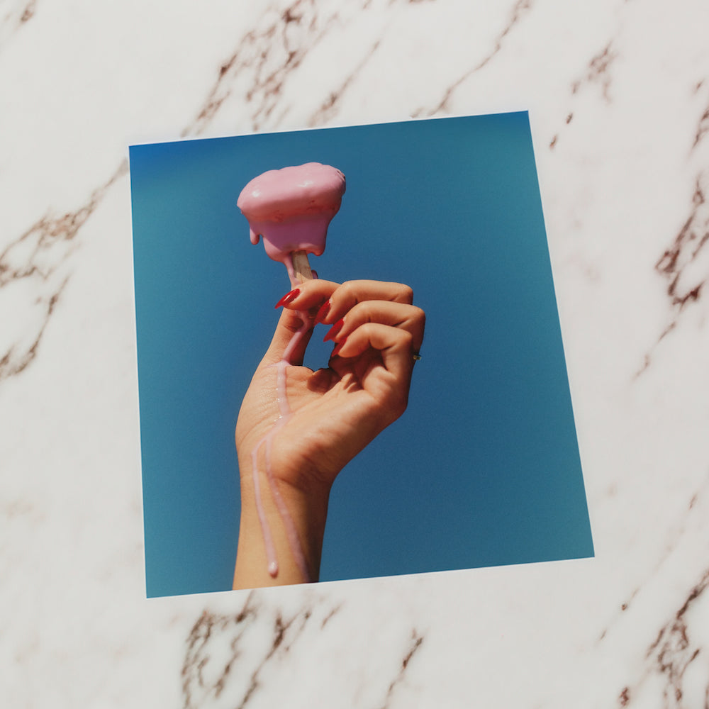 A hand with red nails holding a melting pink ice cream against a blue background.