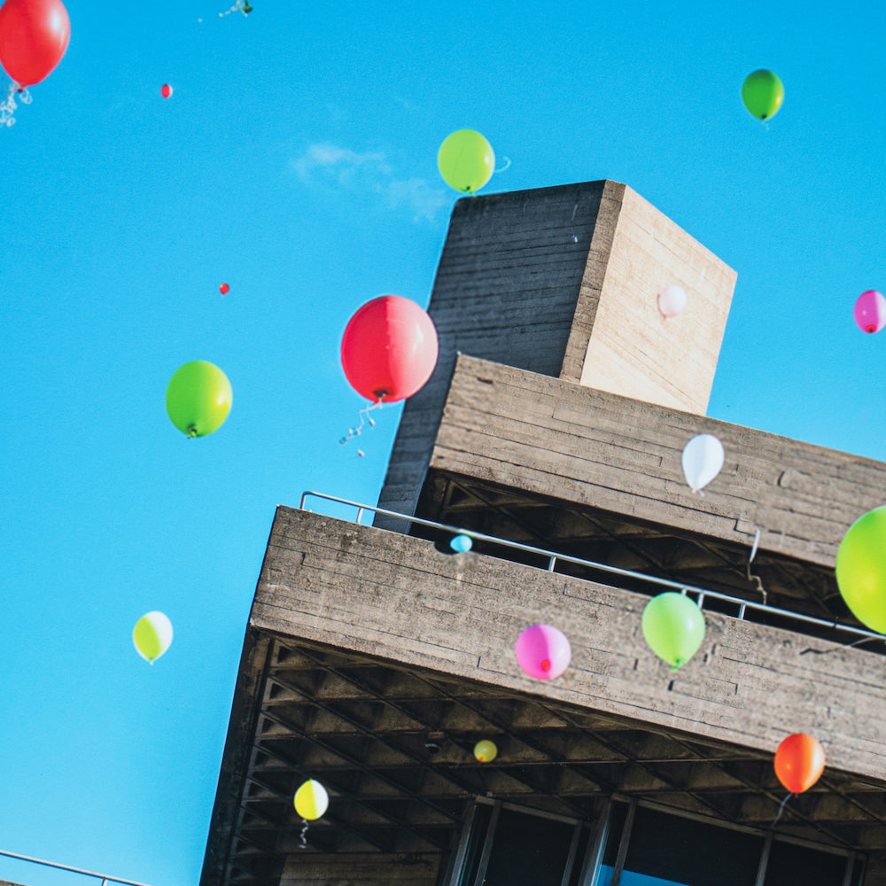 Colorful balloons floating by a concrete building under a clear blue sky.