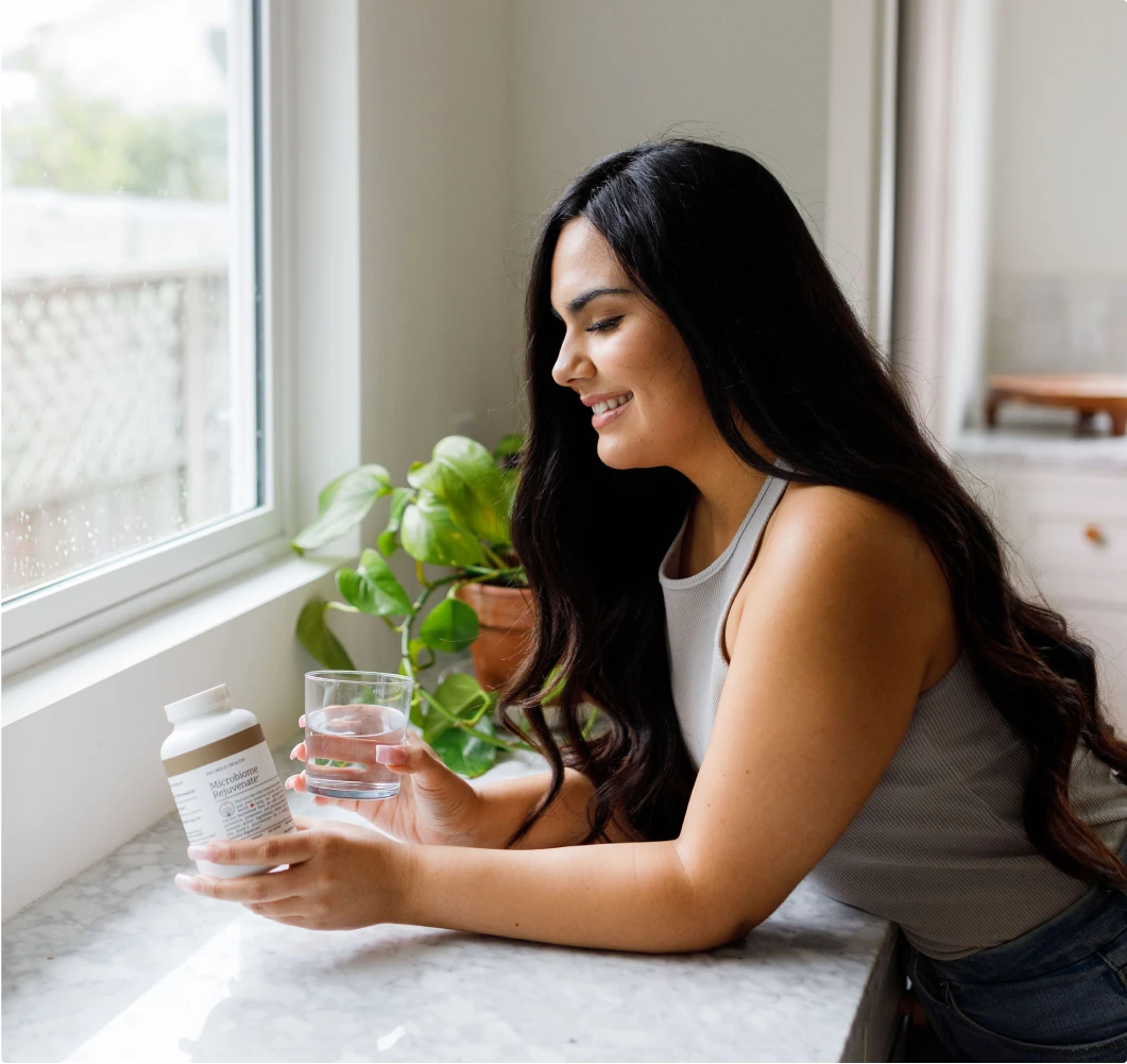 Woman holding a supplement bottle and glass of water, smiling, beside a kitchen window.