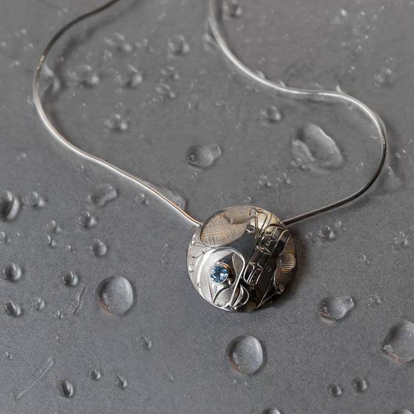 Wolf pendant made with sterling silver. In this depiction by artists Hollie Bartlett, Wolf has large nostrils and prominent fangs emerging from his toothy grin. Hollie uses a blue topaz gemstone for his ovoid eye.
