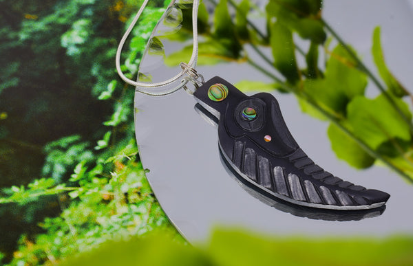 Feather argillite pendant with sharp curve and abalone shell feature. Pendant sits on a mirror with a green forest background, gives the appearance of the pendant floating in water.