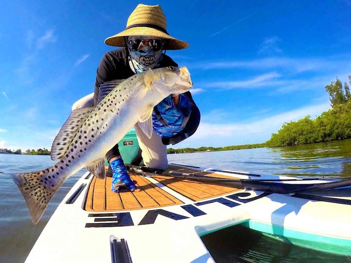 man holding a large fish in front of the camera while riding a paddle board