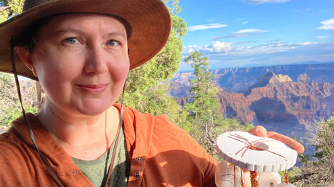 Rebecca Ann Combs is wearing a wide-brimmed brown hat, holding a kumihimo disk in front of the Grand Canyon.