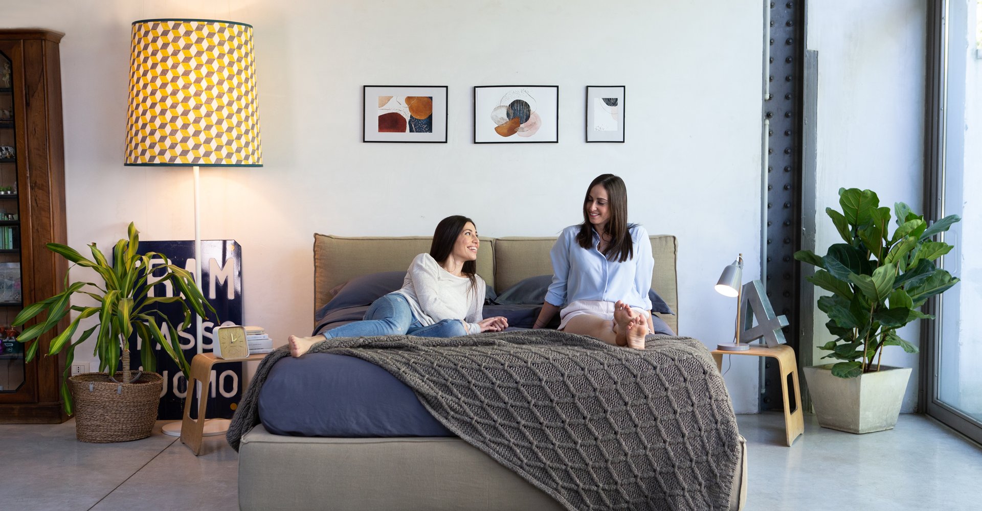 Two middle aged women smiling and talking to each other, as they lay over bed in modern looking bedroom