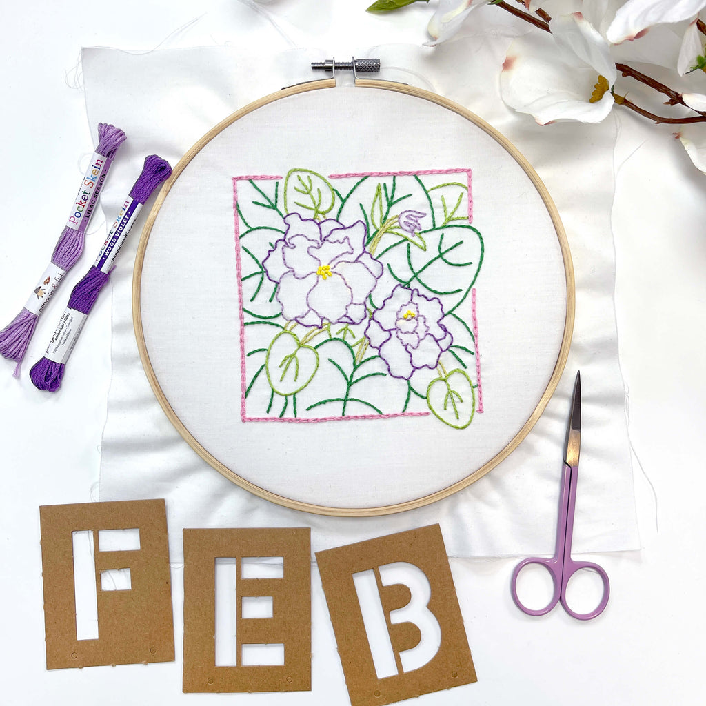 February Violet finished embroidery laid on table with letters F.E.B. under it and a pair of purple scissors