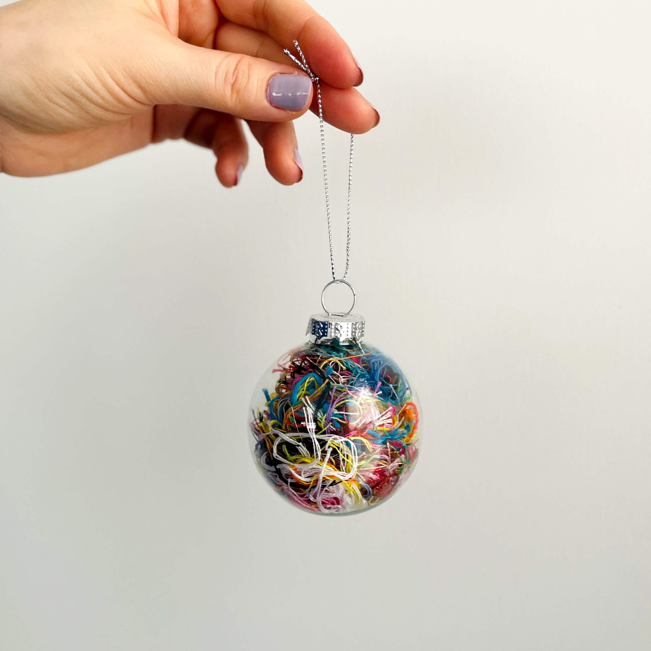 Hand holding a clear christmas ornament filled with scrap embroidery floss in all colors