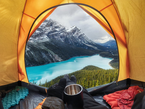 Mensch schaut mit Tasse in der Hand aus einem Zelt auf die Berge