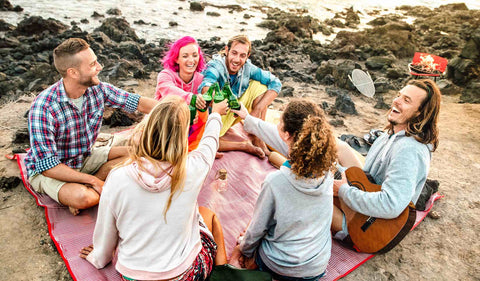 Freunde sitzen auf Decke am Strand und genießen gemeinsam ein kühles Bier.