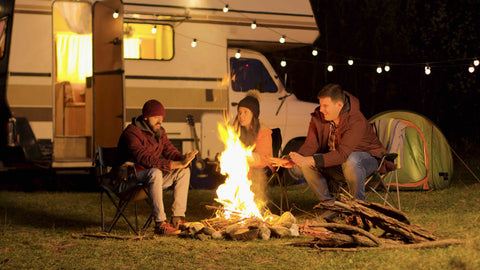 Freunde wärmen sich am Lagerfeuer. Im Hintergrund der Camper und eine Lichterkette. 