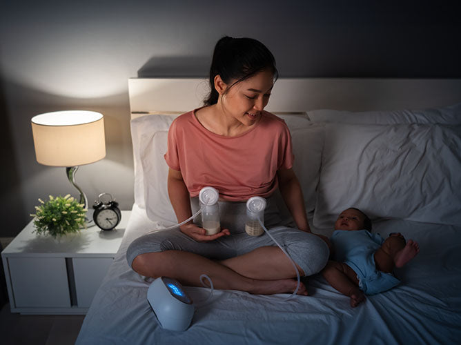 Young mom sitting in bed at night, pumping both of her breasts.