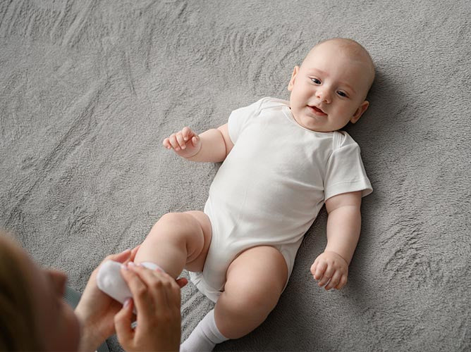 Baby laying in a crib, parents putting on a sock on his right foot.