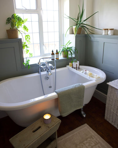 Beautiful ornate stylish bathroom including a bath tub with bath bombs, bath melts and accessories, painted mid green wooden panelled side board complete with plants and Love to b Hand & Body Wash and Hand & Body Lotion.