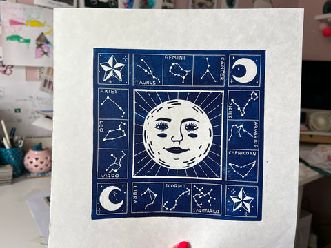 A hand holding a square piece of paper with a blue lino print on it with a sun and star constellations around it