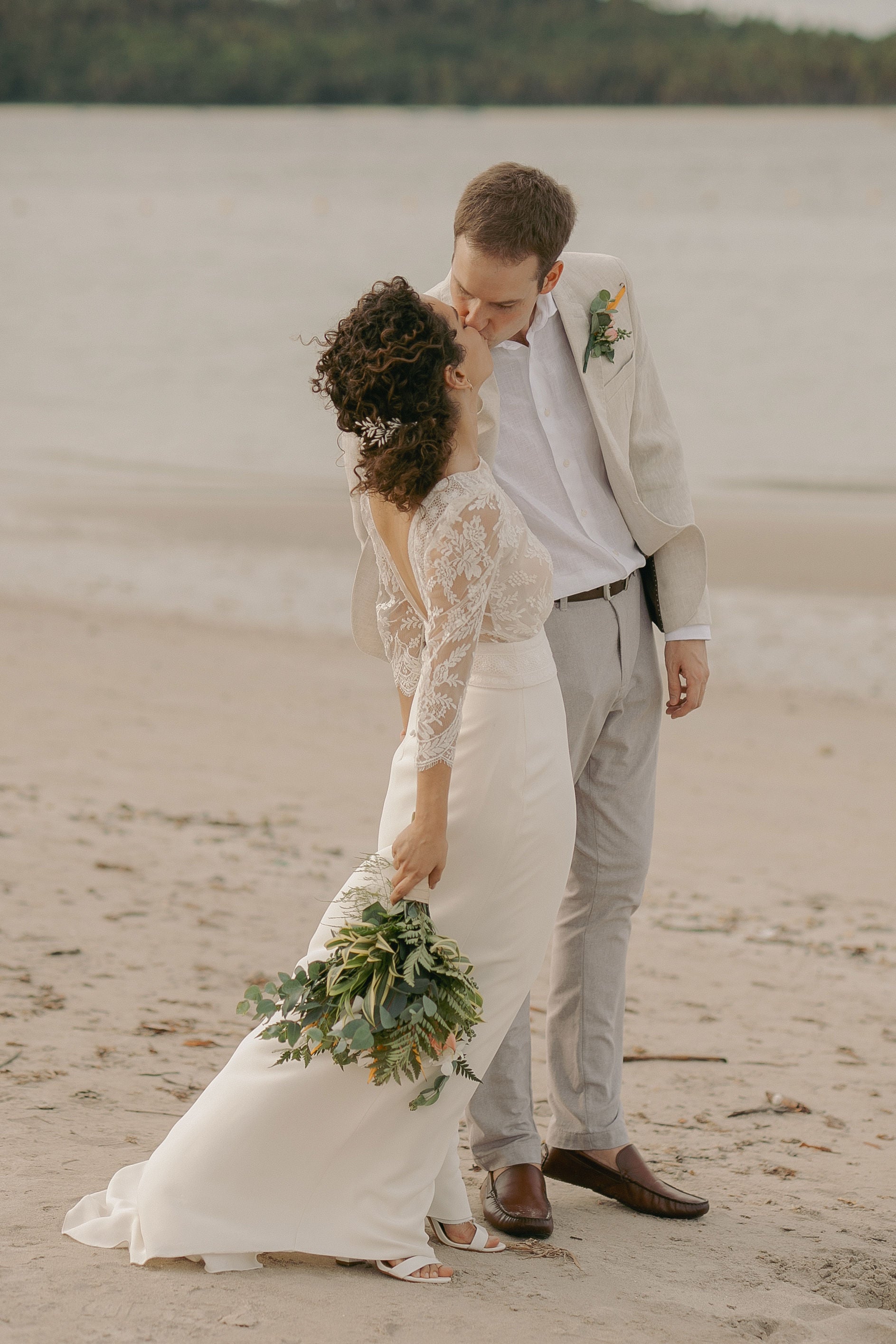 couple de jeunes mariés sur la plage en train de s'embrasser. Robe Laure de Sagazan boucles d'oreilles Lizeron