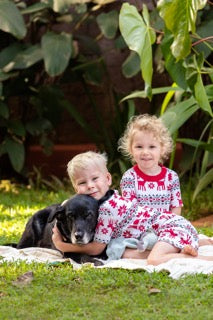 Two children in matching pajamas with a black dog. The background is green tropical foliage.