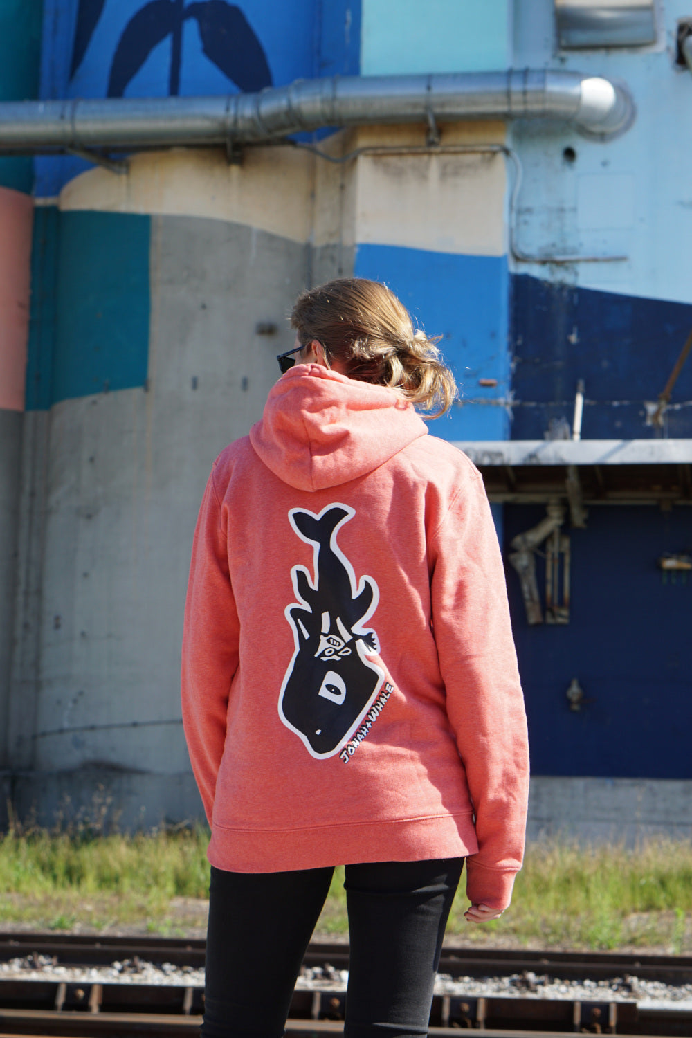 girl wearing a hoodie with a whale logo on back stands in front of a graffiti wall