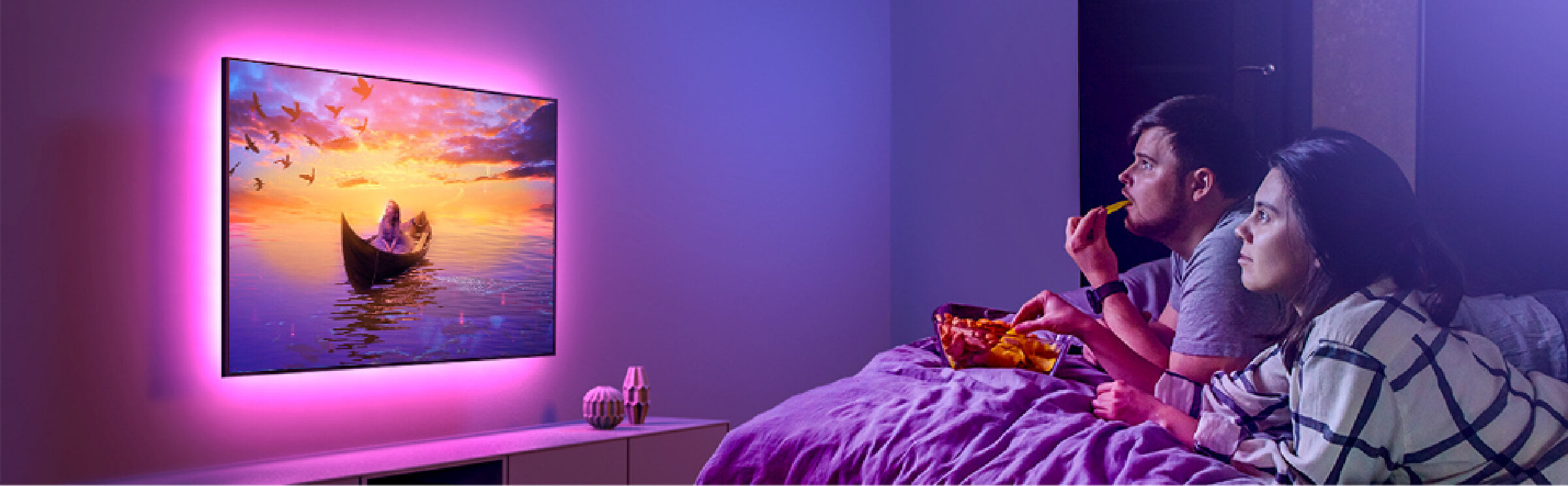 a man and woman sitting on a bed watching a television