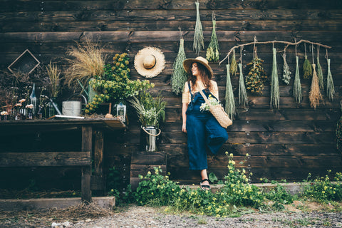 Canadian Tuxedo: woman in denim overalls standing against a barn wall covered in dried flowers. The sun is shining and she is wearing a sunhat. There is a wood table nearby with vases of dried flowers on it. 