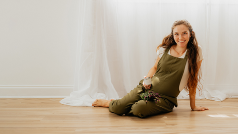 Pregnant woman sitting on the floor, wearing a green jumper, holding a natural stretch mark cream.