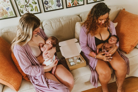 Two women sitting next to each other in purple robes, holding their newborn babies. The one on the left is breastfeeding their baby.