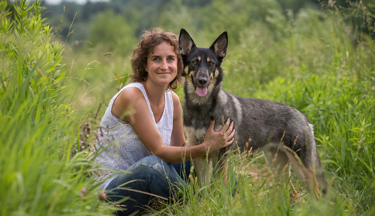 Eine Dame und ein Hund in der Natur blicken glücklich in die Kamera.