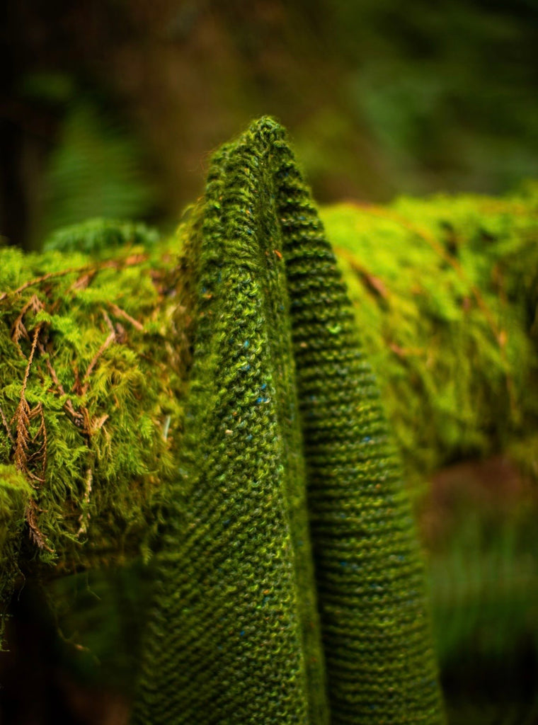 A green garter stitch shawl draped on a mossy log.