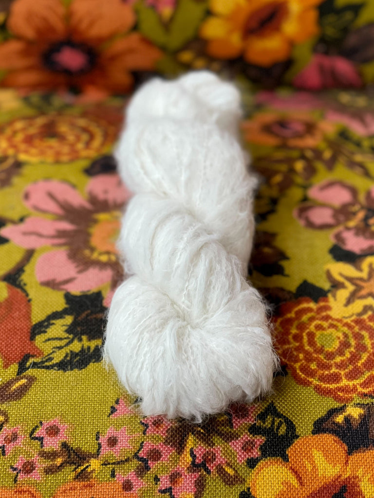 An undyed skein of Dimond Laine Birdie DK yarn laying on a floral background.