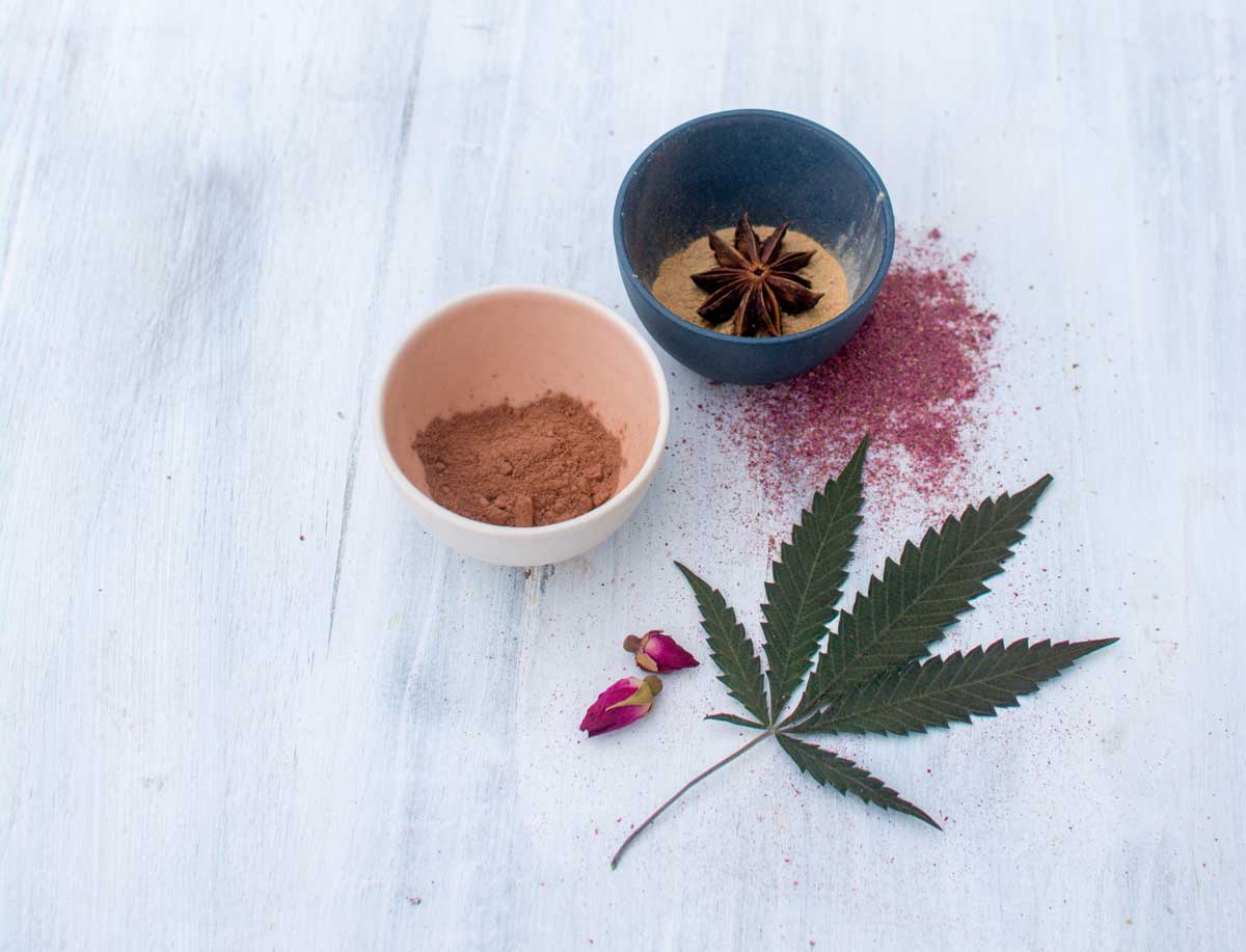 Cocoa powder, rose powder, maca and star anise in small bowls with cannabis leaf for sultry hot cocoa recipe