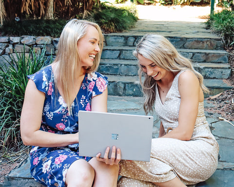 Two women sitting on a step laughing and looking at a laptop computer.