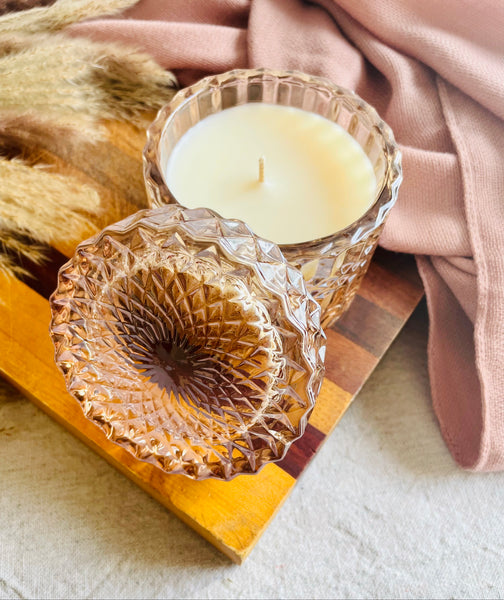 SOi Alluring Amber Shimmer Candle sitting on a wooden board surrounded by a blush scarf and fall twigs